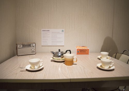 On a linoleum table sits a radio, a box of teabags, a teapot, a milk jug and three teacups