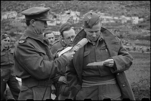 Black and white photo of a soldier reading from a piece of paper into a microphone held by another soldier