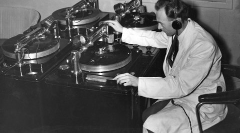 A man sits in front or a large old fashioned recording device. He is wearing a white lab coat and headphones.