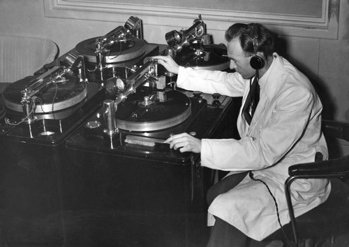 A man sits in front or a large old fashioned recording device. He is wearing a white lab coat and headphones.
