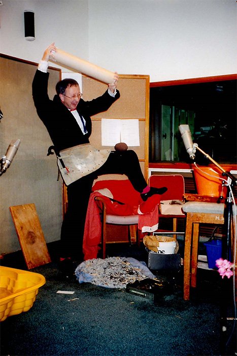 A man in a suit has one leg up on the arm of a chair. He is holding an object above his head and laughing.