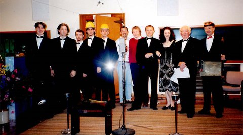 A group of actors stand on a stage in black tie clothes. Two of the men are also wearing yellow hard hats