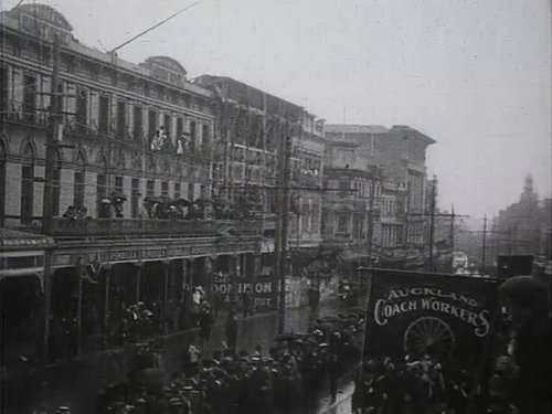 A large crowd of people are gathered in Auckland watching a Labour Day parade. Most are on the street, but there are also people on balconies of buildings holding umbrellas. It is raining.