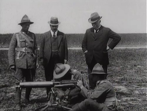 Two men are standing next to two soldiers inspecting a piece of military equipment low to the ground.
