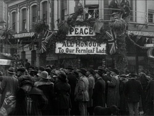 A large crowd stand in front of a building covered in Union Jack flags and a sign that reads ' Peace. All honour to our Fernleaf lads'