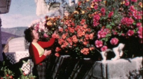 A woman in a red cardigan is standing in her garden with her arms outstretched to some colourful flowers.