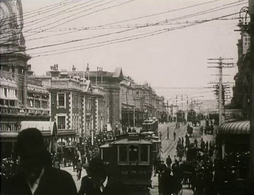 A busy Dunedin street in 1912 filled with people, horse-drawn carts, cars and trams.