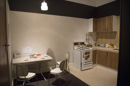 A kitchen, with a linoleum table set for tea, with an oven, bench and cupboards nearby.
