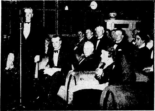 Rt Hon M.J. Savage stands in front of a microphone with several other well-dressed people around him.