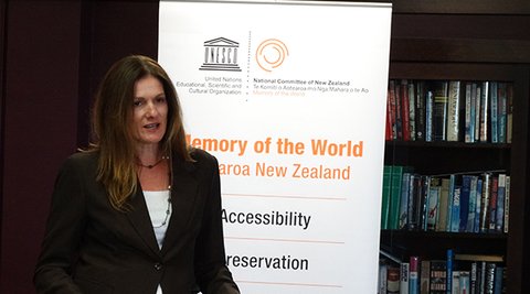 A woman stands in front of a banner for Memory of the World Aotearoa New Zealand