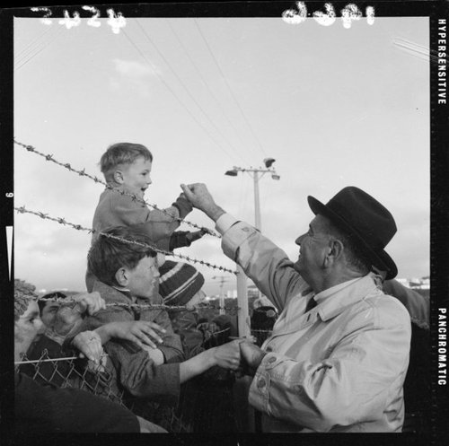 US President Lyndon Johnson reaches over a barbed wire fence to shake the hand of a young boy sitting on his father's shoulders.a
