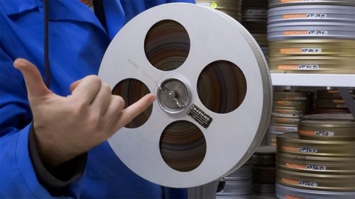 A person in blue safety gear is doing a hand gesture in front of a reel of film.
