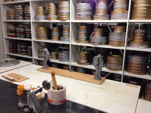 Dozens of neatly stacked film tins on industrial shelves with a film winding bench in the foreground