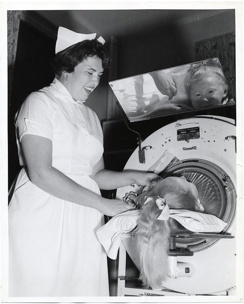 A nurse is reassuring a child who is in an iron lung. The child's face is reflected in a mirror above her.