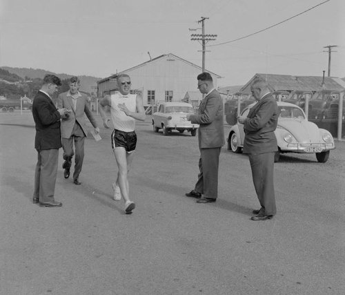 Athlete Norman Read is walking fast, surrounded by other men timing him on watches.