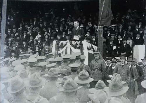 A man gives a speech at a decorated lectern. Behind him are well dressed civilians and facing him are soldiers in WW1 uniforms.