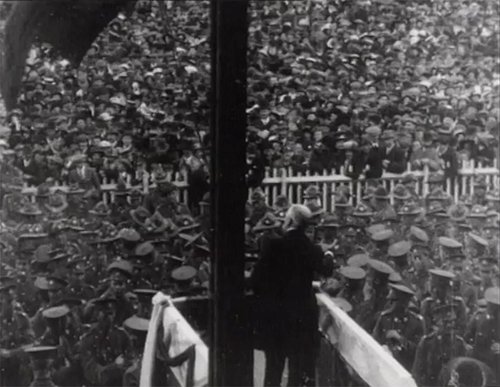 A man stands on a raised platform making a speech in front of a large crowd of soldiers.