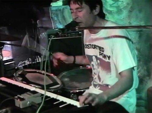 A man is sitting onstage playing the drums and the keyboard and singing into a microphone