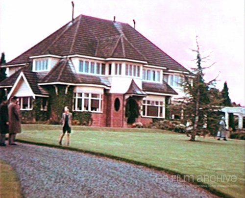 A large house stands in a garden with people outside on the lawn.