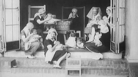 A group of nurses are sitting on a porch feeding babies while other children are nearby.