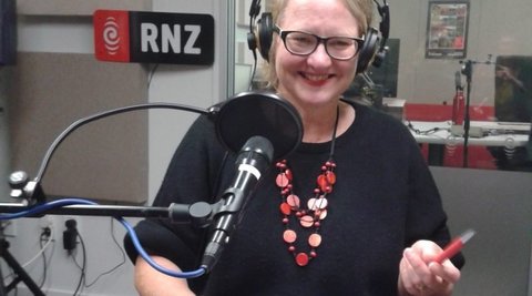 A woman is standing in front of a microphone wearing headphones. She is smiling. Behind her is a sign that reads ' RNZ'