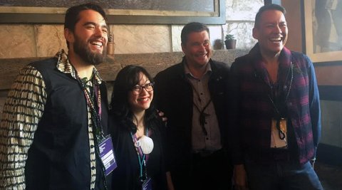 Four people are posing and smiling in front of a wall. They are all wearing lanyards.
