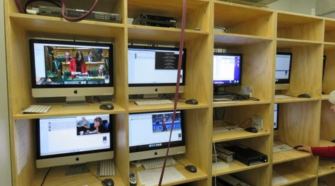 A wooden shelf unit displays many televisions. On one of the screens Jacinda Ardern can be seen speaking in parliament.