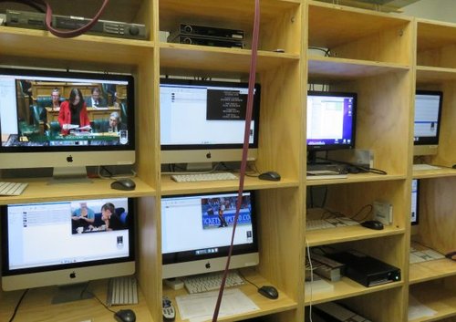 A wooden shelf unit displays many televisions. On one of the screens Jacinda Ardern can be seen speaking in parliament.