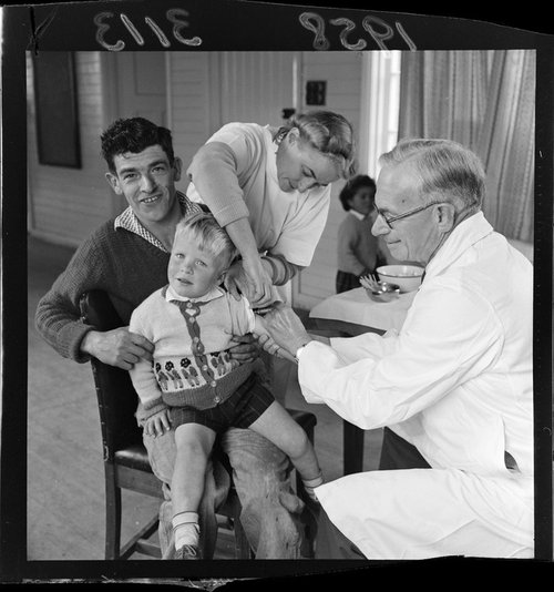 A man holds a child receiving the Polio vaccine from an older male doctor. A female nurse is assisting.