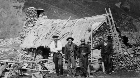 A group of men pose in front of a rough stone hut