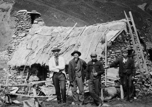 A group of men pose in front of a rough stone hut