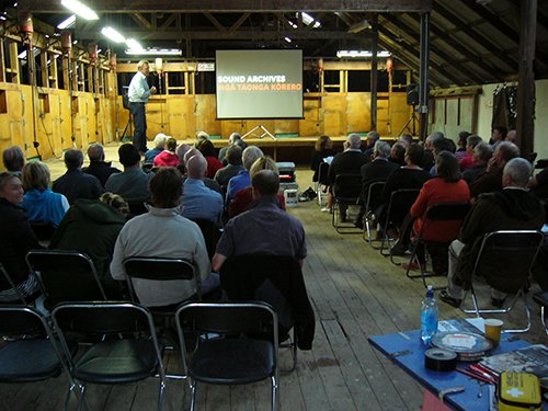 A crowd inside the woolshed are watching a film on a screen.