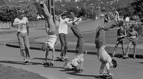 A group of boys are standing around watching three boys riding their skateboards upside down on their hands.