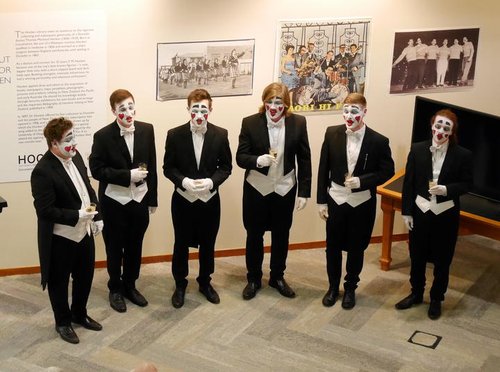 Six mean wearing white and red facepaint are standing in a semi-circle in tuxedos singing.