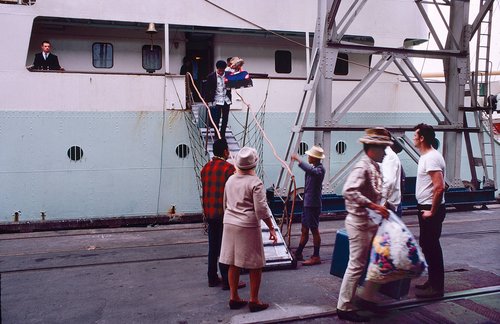 Pacific People disembarking from a boat in Auckland in 1970. They are carrying belongings in suitcases and plastic bags.